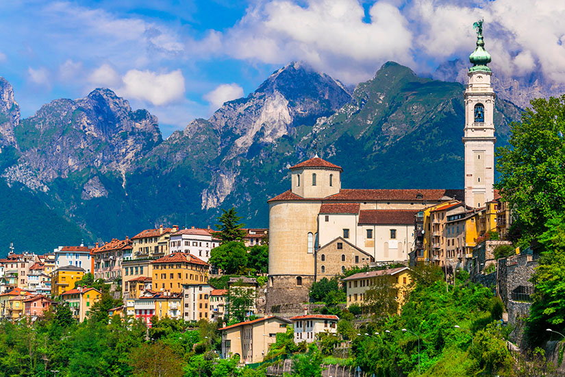 Eine Stadt vor einer Bergkulisse. Im Vordergrund ist eine Kathedrale mit Glockenturm zu sehen. Belluno Stadt