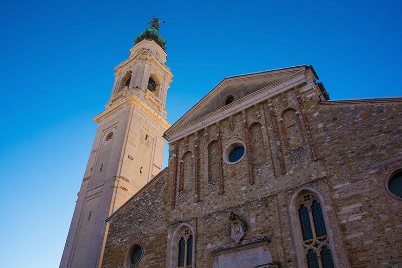 Die Steinfassade einer Kathedrale. Links ist ein hoher Kirchturm zu sehen. Der Himmel ist strahlend blau. Belluno Kathedrale