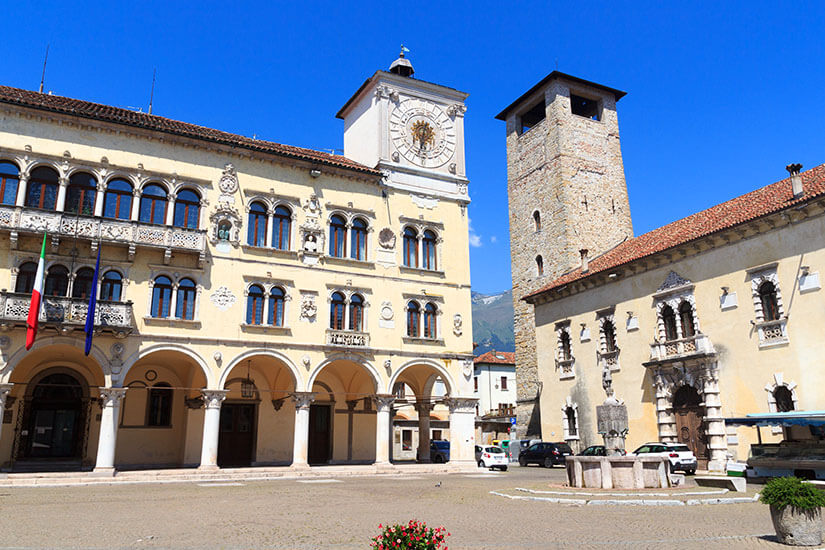 Palast mit Arkadengang und Uhrenturm an einem weitläufigen Platz. Daneben ist ein weiteres Gebäude mit Turm zu sehen. Der Himmel ist blau. Belluno Palazzo dei Rettori