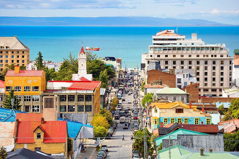Blick über die bunten Dächer einer Stadt. Im Hintergrund ist ein weißer Kirchturm, sowie das Meer zu sehen. Punta Arenas Stadt