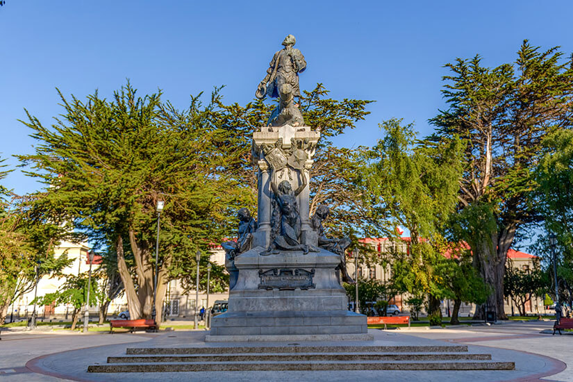 Großes Monument eines Seefahrers, das mitten auf einem Platz steht. Dahinter sind grüne Bäume zu sehen. Punta Arenas Plaza de Armas