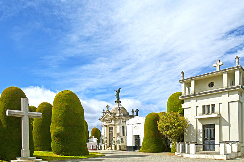 Friedhof mit weißen Mausoleen sowie einem weißen Kreuz. Dazwischen sind grüne Zypressen zu sehen. Punta Arenas Cementerio