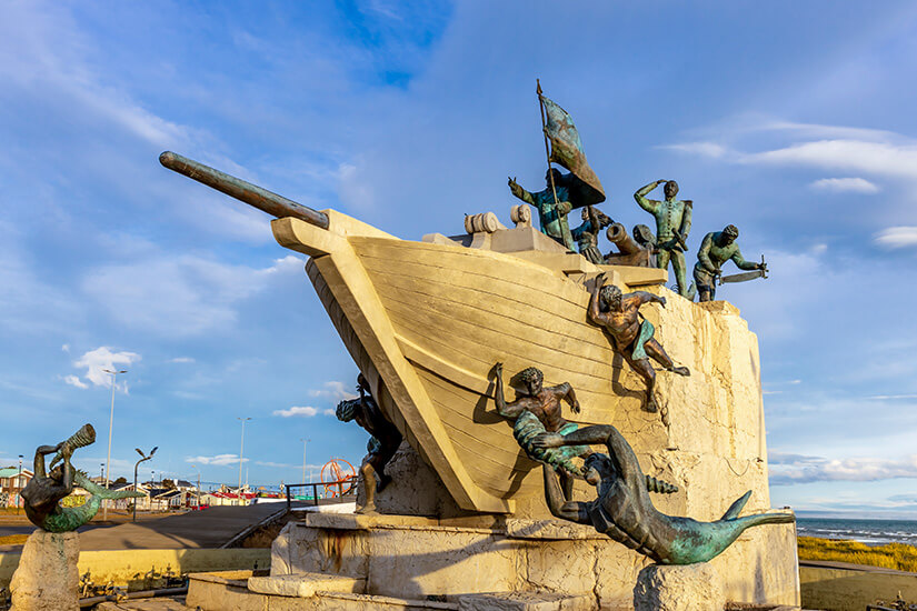 Großes Denkmal in Form eines stilisierten Schiffbugs, an dem mehrere bronzefarbene Figuren in dynamischen Posen hängen oder hinaufklettern. Die Skulptur zeigt Menschen und Meereswesen in einer dramatischen Szene und steht auf einem hellen Sockel unter blauem Himmel. Im Hintergrund sind Küstenlandschaft und Teile einer Promenade zu erkennen. Punta Arenas Museo Nao Victoria