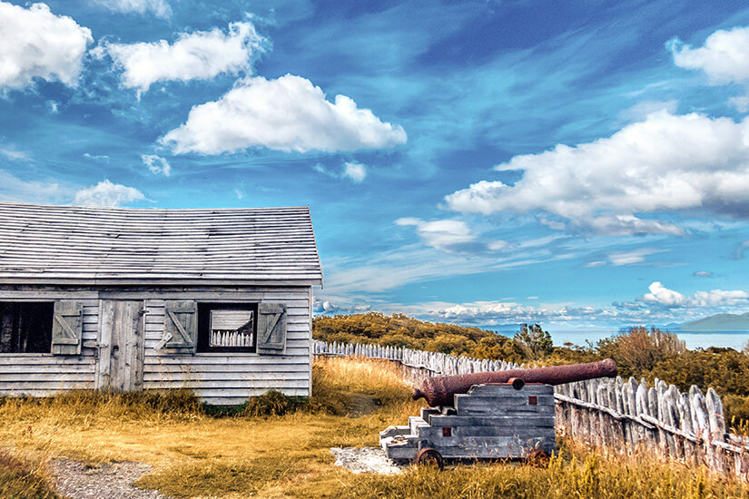 Ein historisches Holzgebäude und eine alte Kanone stehen inmitten einer windgepeitschten Wiesenlandschaft unter einem strahlend blauen Himmel. Ein hölzerner Zaun umrahmt die Szenerie und verstärkt den abgelegenen, patagonischen Charakter des Ortes. Chile Fort Bulnes