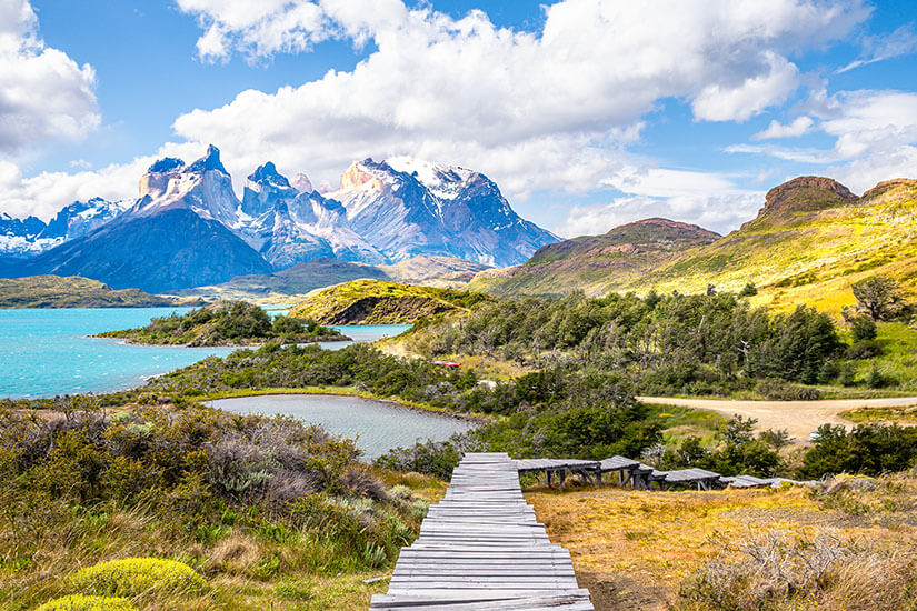 Ein hölzerner Steg führt durch eine farbenfrohe patagonische Landschaft mit türkisblauen Seen und weiten Wiesen. Im Hintergrund erheben sich die markanten, schneebedeckten Berge des Torres-del-Paine-Massivs unter einem teils wolkigen Himmel. Chile Torres del Paine