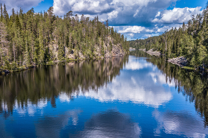 Dichter Nadelwald säumt die felsigen Ufer eines spiegelglatten Sees, in dem sich dramatische weiße Wolken und der blaue Himmel perfekt widerspiegeln. Julma Oelkky Canyonsee