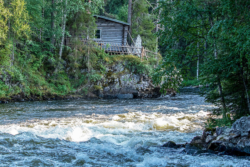 Über einem rauschenden Wildwasserfluss thront eine rustikale Holzhütte auf einem bewachsenen Felsufer, umgeben von dichtem grünem Wald. Kuusamo Myllykoski