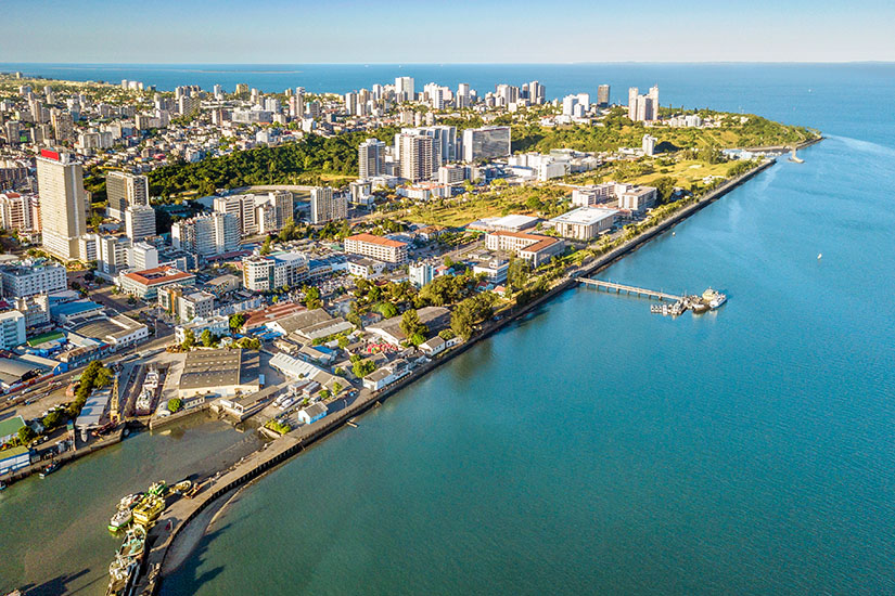 Eine Küstenstadt mit vielen Hochhäusern und dichter Bebauung liegt direkt am Meer. Entlang der Uferlinie verlaufen Straßen und ein langer Pier ragt ins Wasser. Maputo Stadt