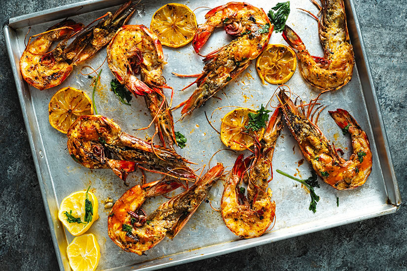 Mehrere gebratene Garnelen liegen gewürzt auf einem Backblech. Dazwischen sind Zitronenscheiben und etwas frische Kräuter verteilt. Mosambik Garnelen