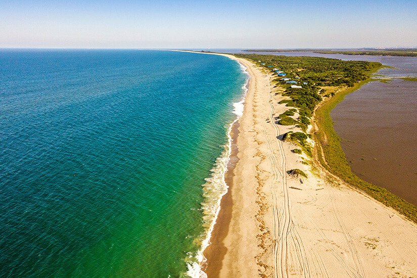 Ein langer, heller Sandstrand zieht sich als schmale Landzunge zwischen Meer und einer Lagune entlang. Links liegt tiefblaues Wasser mit sanften Wellen, rechts ein ruhiger, dunklerer Wasserarm mit Vegetation. Maputo Macaneta