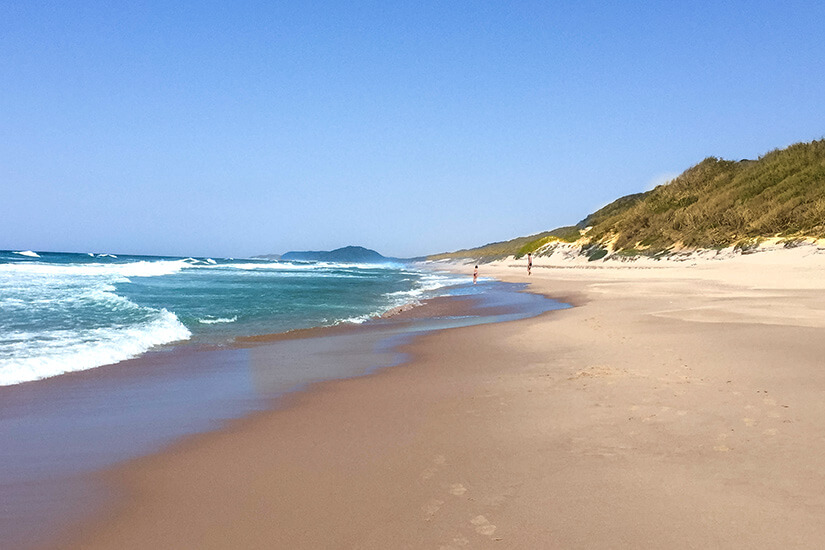 Ein weiter Sandstrand mit sanften Wellen und türkisblauem Meer erstreckt sich bis zum Horizont. Rechts liegen Dünen und bewachsene Hügel, links öffnet sich das offene Wasser. In der Ferne sind ein paar Personen am Strand und eine kleine Landzunge zu erkennen. Mosambik Ponta Do Ouro