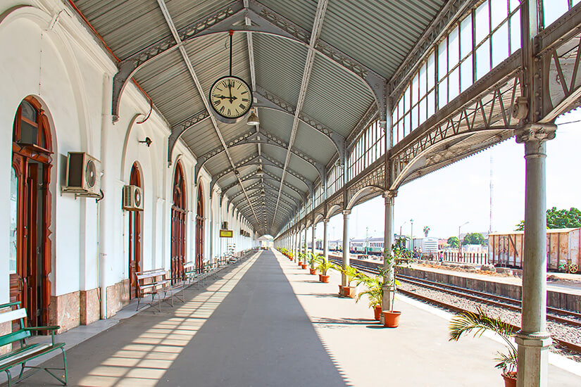 Ein langer, überdachter Bahnsteig mit Stahlkonstruktion und großen Fenstern zieht sich in die Tiefe. Links stehen historische Gebäudefassaden mit Bögen, Bänken und einer großen Uhr hängt unter dem Dach. Rechts verlaufen Gleise, daneben stehen mehrere Pflanzkübel entlang des Bahnsteigs. Maputo Bahnhof
