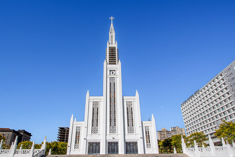 Eine große, weiß gestrichene Kathedrale mit sehr hoher, schlanker Turmspitze und einem Kreuz oben. Die Fassade ist streng symmetrisch mit hohen, schmalen Fenstern und geometrischen Details. Maputo Kathedrale