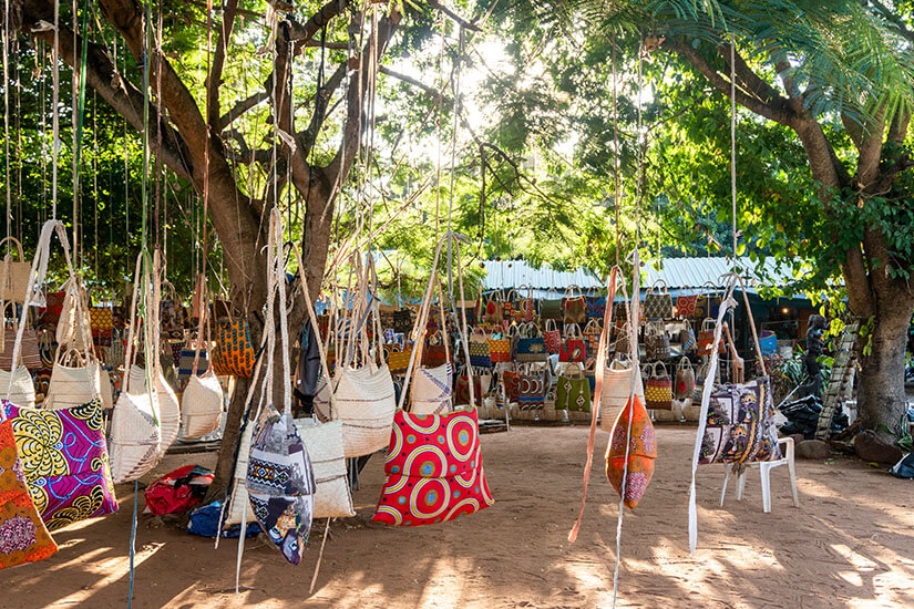 Ein Marktstand im Schatten großer Bäume, an dem viele bunte Taschen und Stoffbeutel an Schnüren hängen. Im Hintergrund sind weitere Stände mit Textilien und Handwerkswaren zu sehen, dazu ein paar Stühle und Gegenstände auf dem sandigen Boden. Maputo FEIMA