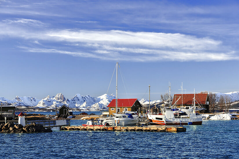 Ein Ortsteil der Stadt Sortland in Norwegen ist zu sehen. Häuser mit roten Dächern stehen direkt am Wasser, weiße Boote liegen davor, im Hintergrund sind verschneite Berge zu sehen. Vesteralen Norwegen Sortland
