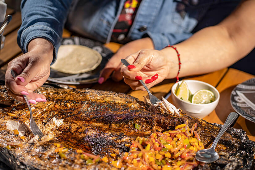 Mehrere Hände mit bunt lackierten Nägeln zupfen mit Gabeln saftige Stücke von einem großen, knusprig gegrillten Fisch auf einem Holzbrett. Chilenisches Essen Pescado
