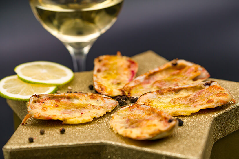 Goldbraun überbackene Jakobsmuscheln präsentieren sich elegant auf einem bronzefarbenen Teller, garniert mit Limetten- und Zitronenscheiben sowie Pfefferkörnern. Machas a la Parmesana