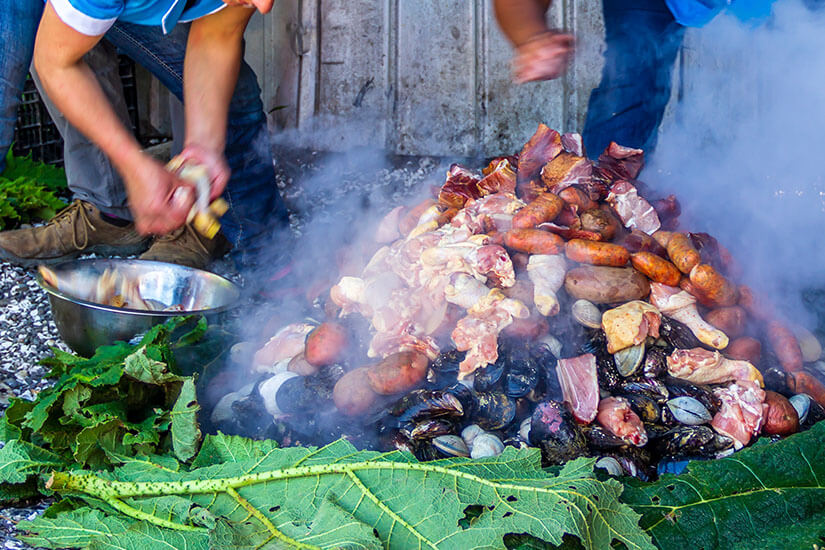 Dampfende Schichten von Fleischstücken, Würstchen, Hähnchen, Muscheln, Venusmuscheln und Kartoffeln türmen sich auf, während dichter Rauch aufsteigt. Chilenisches Essen Curanto