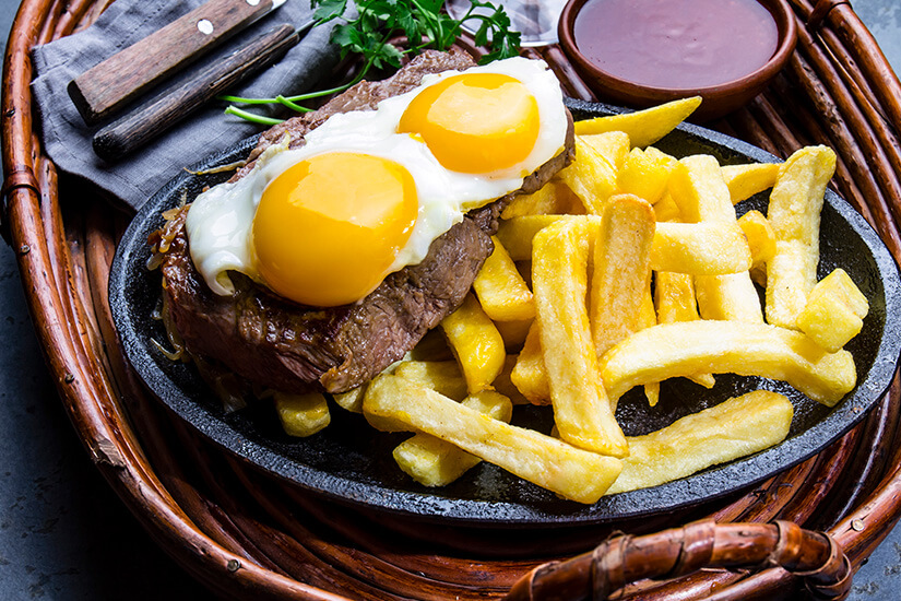 Saftiges Rindersteak, gekrönt von zwei Spiegeleiern mit leuchtend gelben Dottern, brutzelt auf einer heißen Steinplatte neben goldgelben, dicken Pommes frites. Chilenisches Essen Rinderfilet