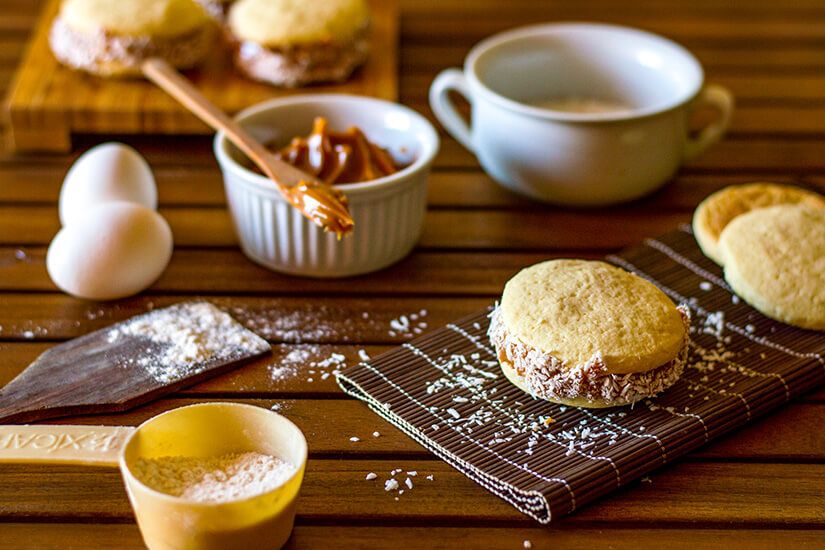 Zarte, mürbe Doppelkekse gefüllt mit cremigem Dulce de Leche und am Rand mit Kokosraspeln verziert liegen auf einer Bambusmatte, umgeben von den Backzutaten. Chilenisches Essen Alfajores