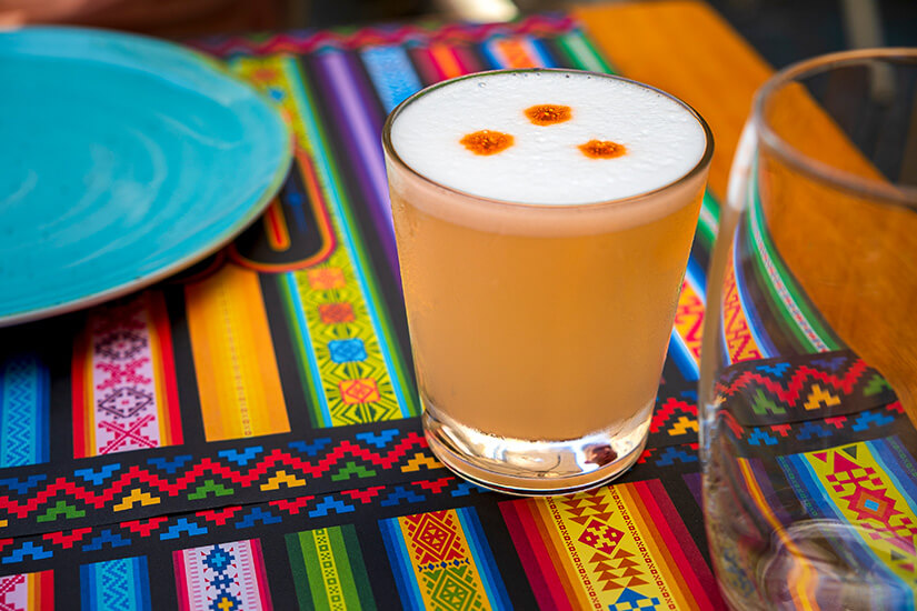 Schaumgekrönter Pisco Sour mit zwei Tropfen Angostura-Bitter auf der weißen Eiweißhaube schimmert goldgelb im Glas auf einem farbenfrohen, traditionell andinen Tischläufer. Chilenisches Essen Pisco Sour