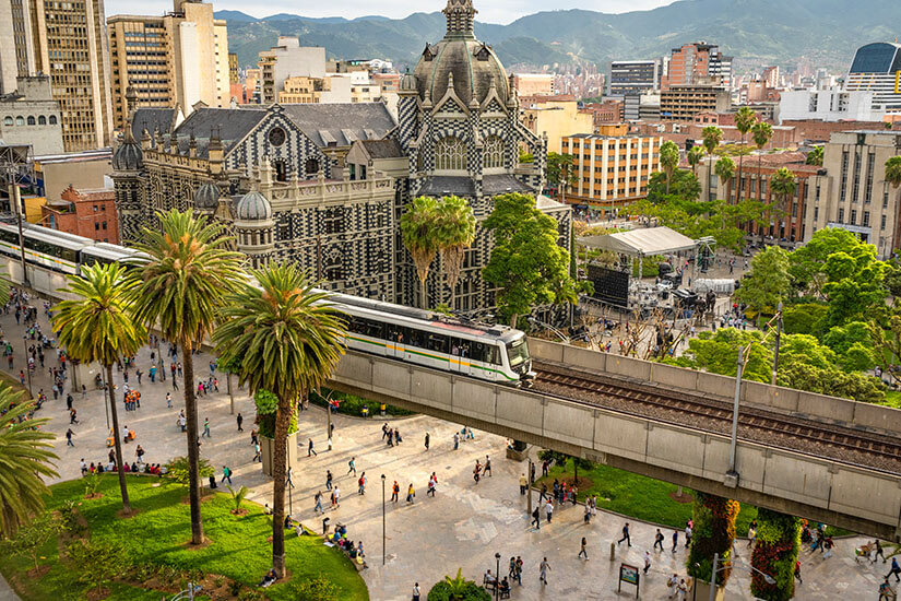 Blick von oben auf die belebte Plaza Botero in Medellín, Kolumbien. Eine moderne Metro-Bahn passiert den historischen Palacio de la Cultura Rafael Uribe Uribe inmitten von grünen Palmen und Stadtbesuchern. Medellín Kolumbien Willkommen
