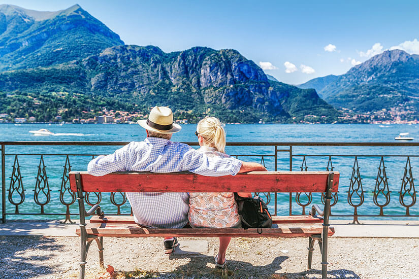 Romantisches Paar sitzt eng umschlungen auf einer roten Holzbank an einer Uferpromenade und genießt den Blick über das tiefblaue Wasser auf die dramatischen Berge am gegenüberliegenden Ufer. Fruehbucher oder Lastminute Paar