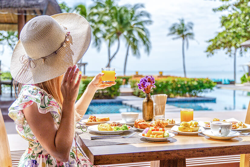 Eine Frau mit großem Strohhut und geblümtem Kleid genießt ein üppiges Frühstück mit frischen Früchten, Gebäck, Eiern und Orangensaft, während sie den Blick über den Pool auf das Meer und die Palmen schweifen lässt. Fruehbucher oder Lastminute Verpflegung