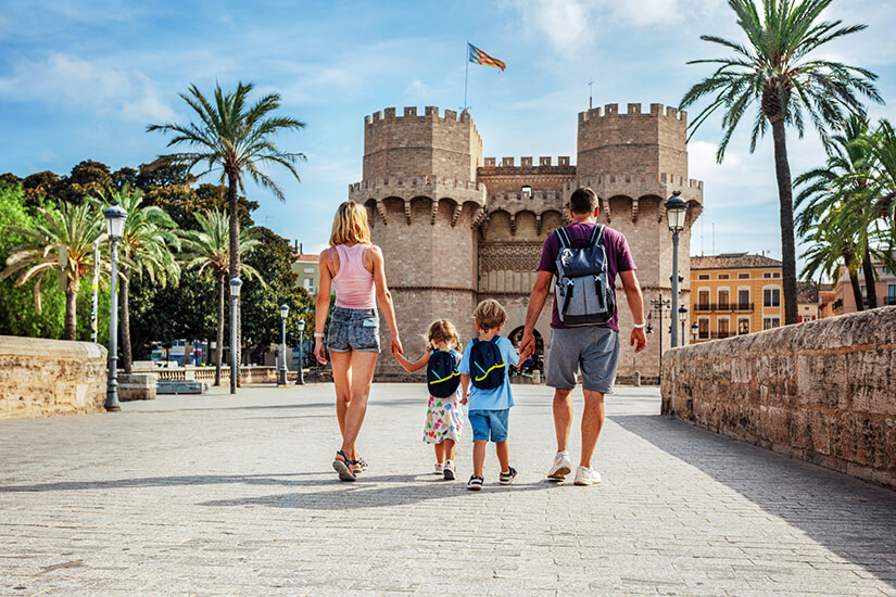 Mutter und Vater mit zwei kleinen Kindern spazieren Hand in Hand über eine breite Steinbrücke auf ein imposantes mittelalterliches Stadttor mit zwei mächtigen Wehrtürmen zu, flankiert von Palmen und einer wehenden Flagge. Fruehbucher oder Lastminute Familie