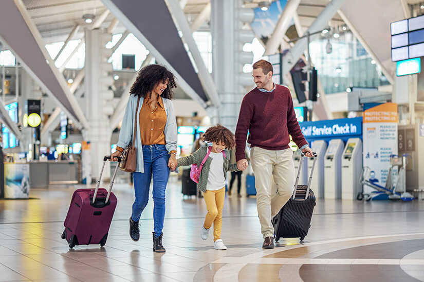 Eine fröhliche Familie – Mutter, Vater und ein kleines Kind mit Locken – schreitet strahlend durch eine moderne Flughafenhalle, während das Kind ausgelassen zwischen den Eltern hüpft und beide Hände hält. Fruehbucher oder Lastminute Flughafen
