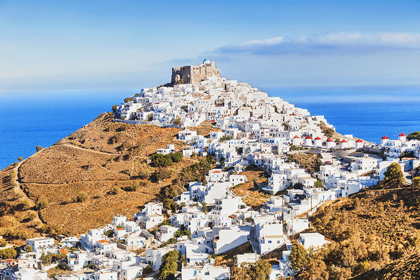 Eine dicht gebaute Siedlung aus weißen Häusern zieht sich einen Hügel hinauf bis zu einer Festung auf der Spitze. Rundherum liegt karges, braunes Gelände, dahinter erstreckt sich das tiefblaue Meer. Astypalea Stadt