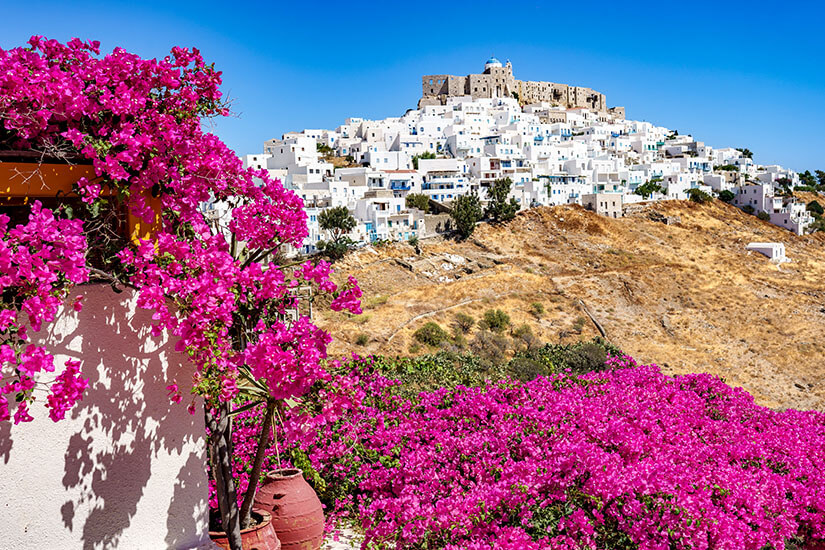 Leuchtend pinke Bougainvillea-Blüten füllen den Vordergrund und rahmen einen Hügel mit dicht gedrängten weißen Häusern ein. Oben thront eine steinerne Festung über der Siedlung. Astypalea Sommer