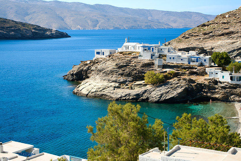 Weiß getünchte Häuser schmiegen sich an eine felsige Landzunge, die ins tiefblaue Meer der Ägäis ragt. Im Vordergrund rahmen Pinien das Bild. Im im Hintergrund sind die kargen Berge einer Nachbarinsel zu sehen. Tinos Griechenland Dorf