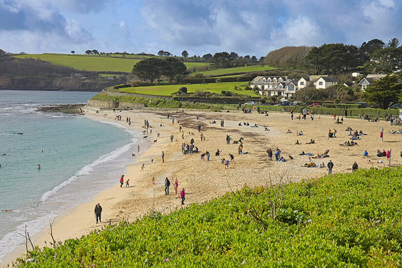 Zahlreiche Strandbesucher tummeln sich auf einem goldenen Sandstrand vor einer Kulisse aus saftig grünen Weiden, weißen Cottages und sanften Hügeln unter einem wechselhaften Himmel. Cornwall Straende Gyllyngvase Beach