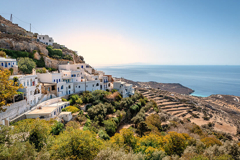 Ein traditionelles Bergdorf mit weiß getünchten Häusern und blauen Fensterläden schmiegt sich an einen felsigen Hang. Olivenbäume und üppige Vegetation umgeben die terrassenförmig angelegten Gebäude. Im Hintergrund erstreckt sich der Blick über landwirtschaftliche Terrassen bis zur ruhigen Ägäis, am Horizont zeichnet sich die Silhouette einer Nachbarinsel ab. Tinos Griechenland Kardiani