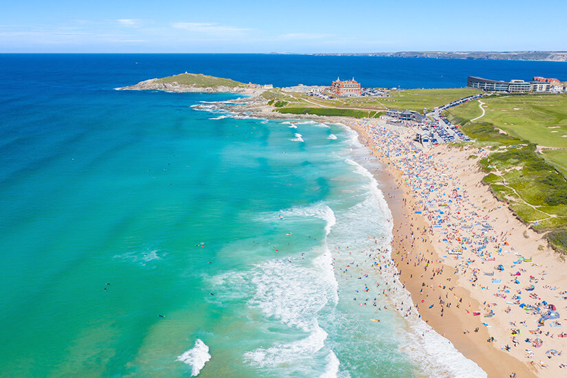 Spektakuläre Luftaufnahme eines dicht belegten Strandes mit türkisfarbenem Wasser und perfekten Surfwellen, während sich bunte Sonnenschirme und Badegäste auf dem goldenen Sand drängen. Cornwall Straende Fistral