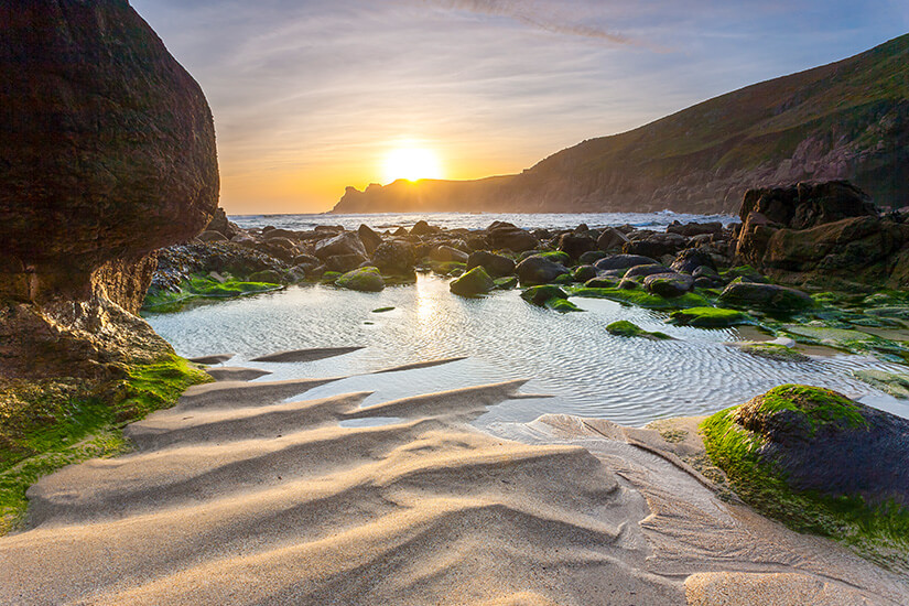 Abendlicht flutet eine versteckte Felsenbucht, in der sich Gezeitentümpel zwischen moosbewachsenen Steinen und kunstvoll vom Wasser geformten Sandrippen sammeln. Cornwall Straende Nanjizal Bay