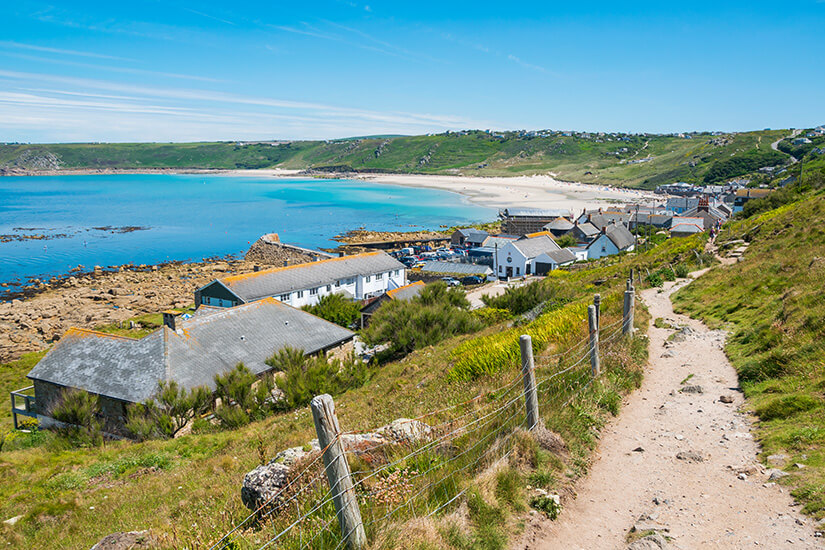 Ein Sandiger Küstenpfad schlängelt sich einen grünen Hang hinab zu einem idyllischen Fischerdorf mit weißen Cottages und Schieferdächern, während dahinter eine geschwungene Bucht mit türkisblauem Wasser und hellem Sandstrand lockt. Cornwall Straende Sennen Cove