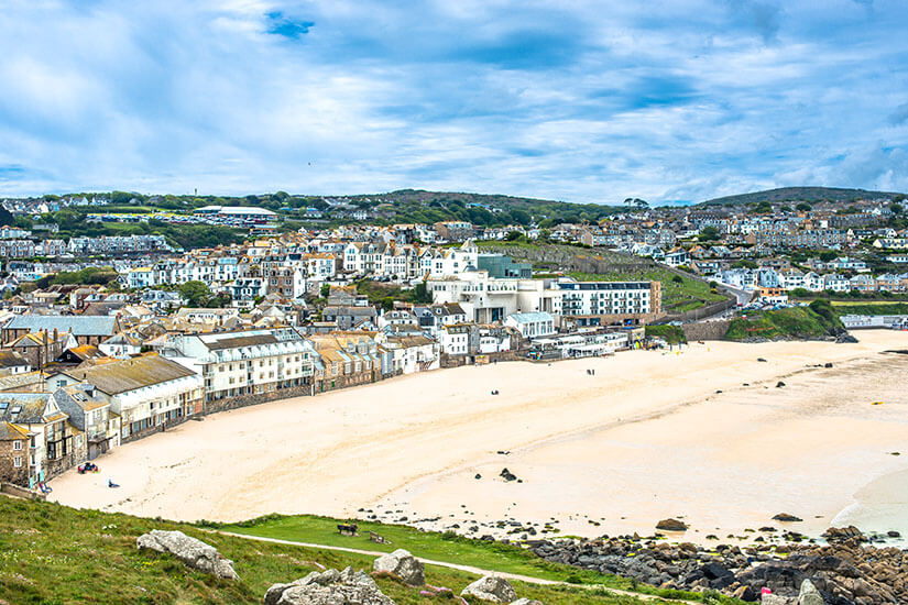 Panoramablick über einen weitläufigen Sandstrand und eine malerische Küstenstadt mit dicht gedrängten weißen und pastellfarbenen Häusern, die sich terrassenförmig den Hang hinaufziehen. St Ives Porthmeor Beach