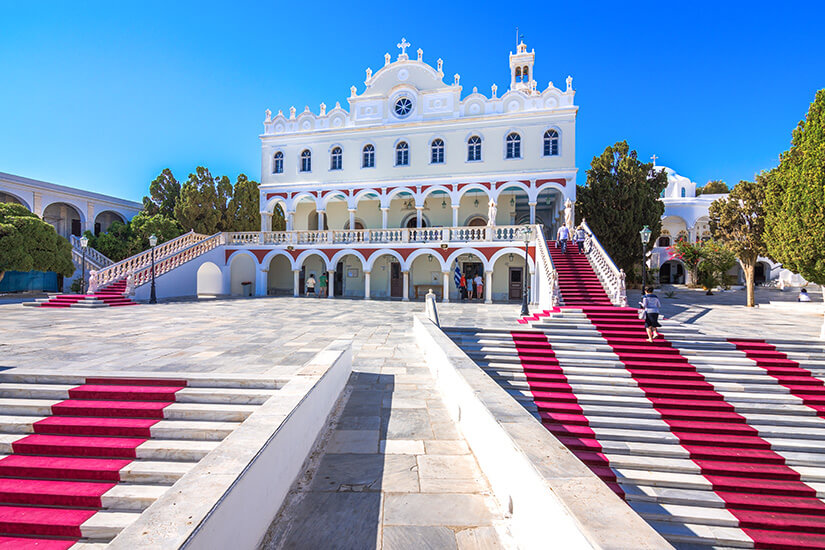 Die imposante Wallfahrtskirche Panagia Evangelistria thront auf einem Hügel über Tinos Stadt. Breite Marmortreppen mit roten Teppichen führen hinauf zum weiß getünchten Gebäude mit seinen eleganten Arkaden und Balkonen. Der weitläufige Vorplatz aus poliertem Marmor glänzt unter dem strahlend blauen Himmel. Tinos Griechenland Panagia Megalochari Evangelistria