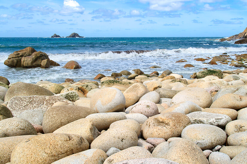 Glatt geschliffene Granitfelsen in warmen Beige- und Rosatönen bedecken den Strand, während Wellen gegen die Steine branden. Cornwall Straende Porth Nanven
