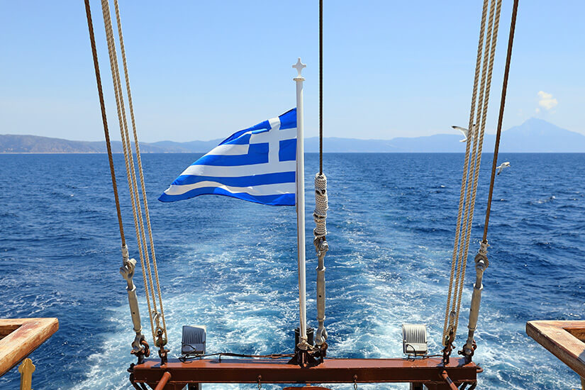 Vom Heck eines Boots aus schweift der Blick über die Ägäis. Die griechische Flagge flattert im Wind. Am Horizont zeichnen sich die Silhouetten der Kykladeninseln ab. Tinos Griechenland Boot
