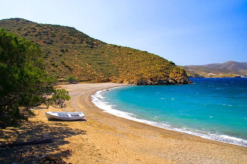 Ein ruhiger, geschwungener Strand mit hellem Kies und türkisfarbenem Wasser liegt in einer kleinen Bucht. Am Ufer steht ein einzelnes Boot im Schatten eines Baumes. Die umliegenden Hügel sind trocken und spärlich bewachsen. Astypalea Livadi Beach