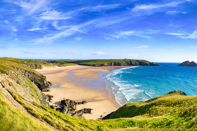 Eine geschwungene Bucht mit goldenem Sand, eingerahmt von sattgrünen Klippen und Landzungen, während sanfte Wellen türkisfarbenes Wasser an den Strand spülen und kleine Felseninseln am Horizont auftauchen. Cornwall Straende Holywell Bay