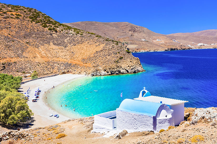 Eine kleine, türkisfarbene Bucht liegt zwischen kargen Hügeln mit einem hellen Kiesstrand. Im Vordergrund steht eine weiße Kapelle mit blauem Dach und Kreuz. Am Strand sind einige Badende und Sonnenschirme zu erkennen, das Meer wirkt sehr klar. Astypalea Agios Konstantinos Beach