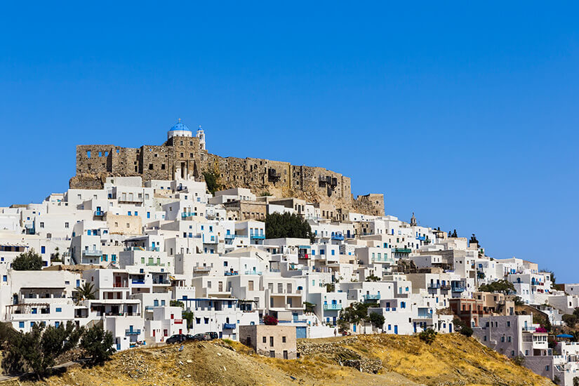 Weiße, würfelförmige Häuser ziehen sich dicht an einem Hügel hinauf. Ganz oben thront eine massive steinerne Burganlage mit einer kleinen Kirche. Der Himmel ist klar und intensiv blau. Astypalea Burg