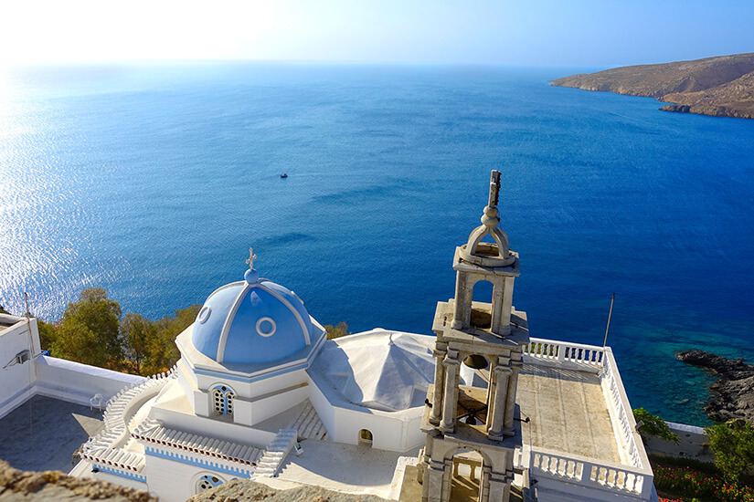 Eine weiß-blaue Kirche mit Kuppel und einem Glockenturm liegt hoch über dem tiefblauen Meer. Terrassen und Balustraden umgeben das Gebäude und öffnen den Blick bis zum Horizont. Astypalea Kirche