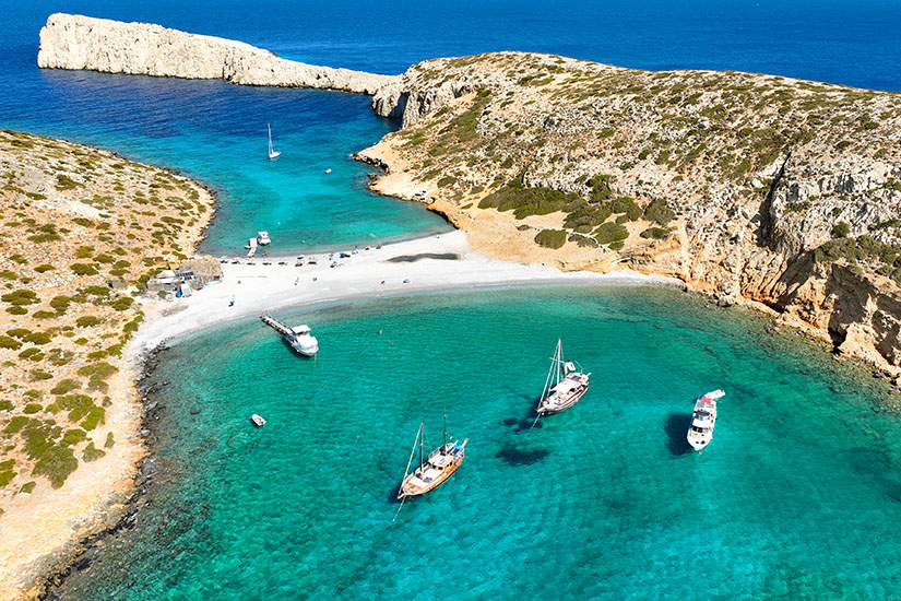 Eine kleine Sandbank verbindet zwei felsige Landzungen und teilt eine türkisfarbene Lagune. Mehrere Segel- und Motorboote ankern im klaren Wasser. Griechenland Bootsfahrt