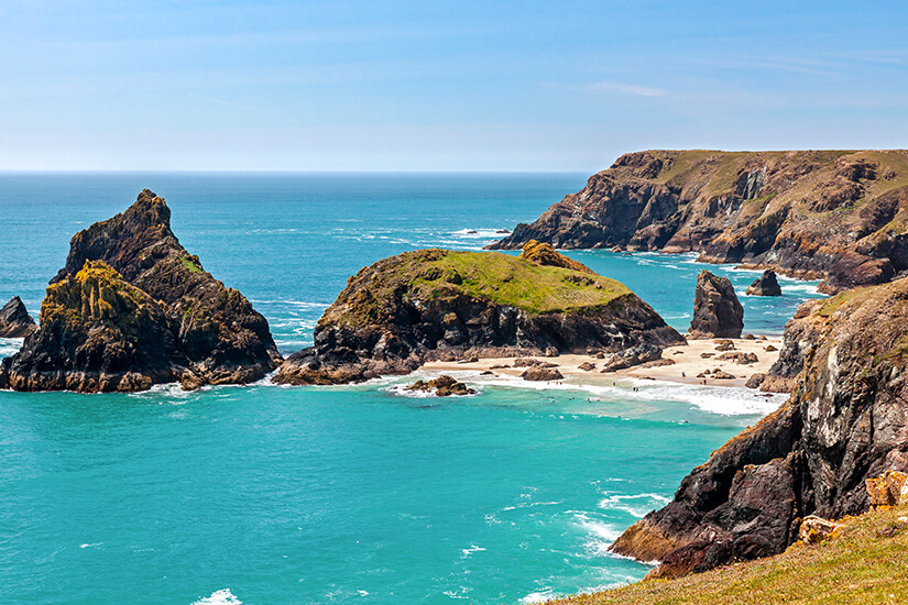 Dramatische Felsnadeln und grün bewachsene Inselchen ragen aus dem türkisfarbenen Wasser einer zerklüfteten Bucht, während sich im Hintergrund schroffe Klippen entlang der Küste erstrecken. Kynance Cove