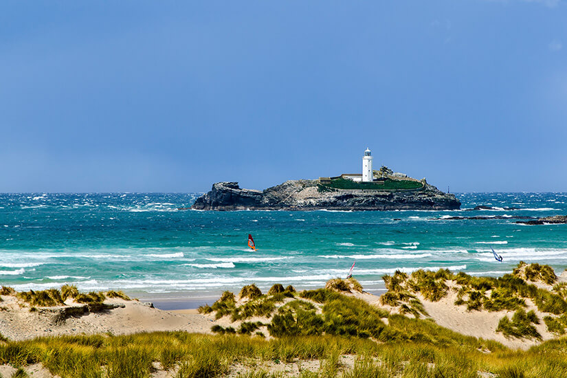 Ein weißer Leuchtturm thront auf einer kleinen Felseninsel vor türkisfarbener, aufgewühlter See, während Windsurfer durch die Wellen gleiten und Sanddünen mit Strandhafer den Vordergrund bilden. Gwithian Towans